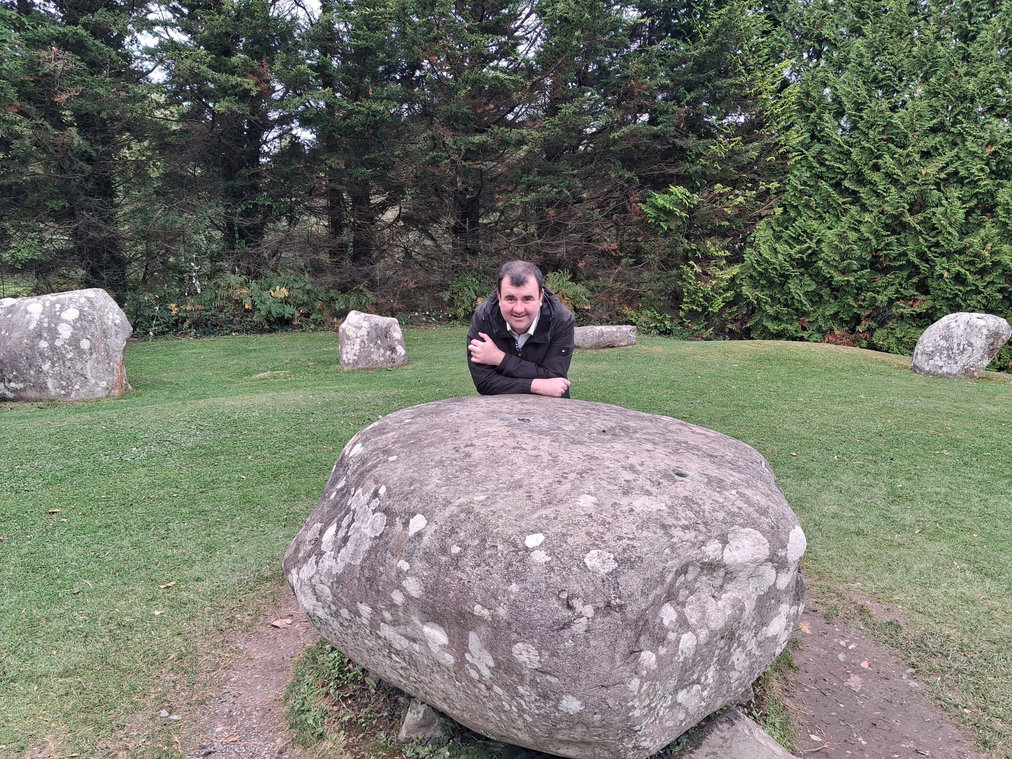 Paul leans on a large boulder in the middle of a stone circle, with tall trees behind.