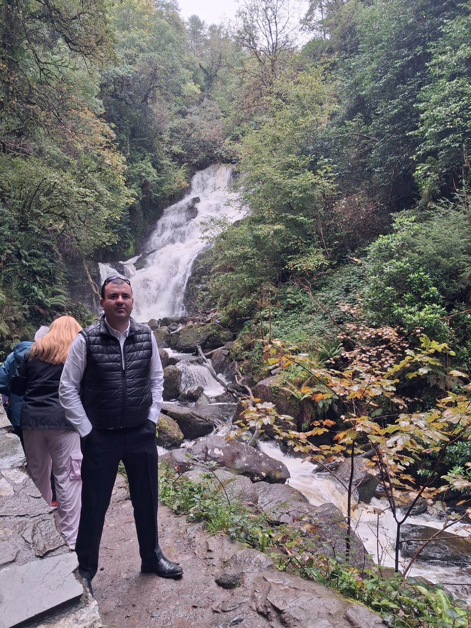Paul stands on a path beside a waterfall that runs down through a wooded glen.