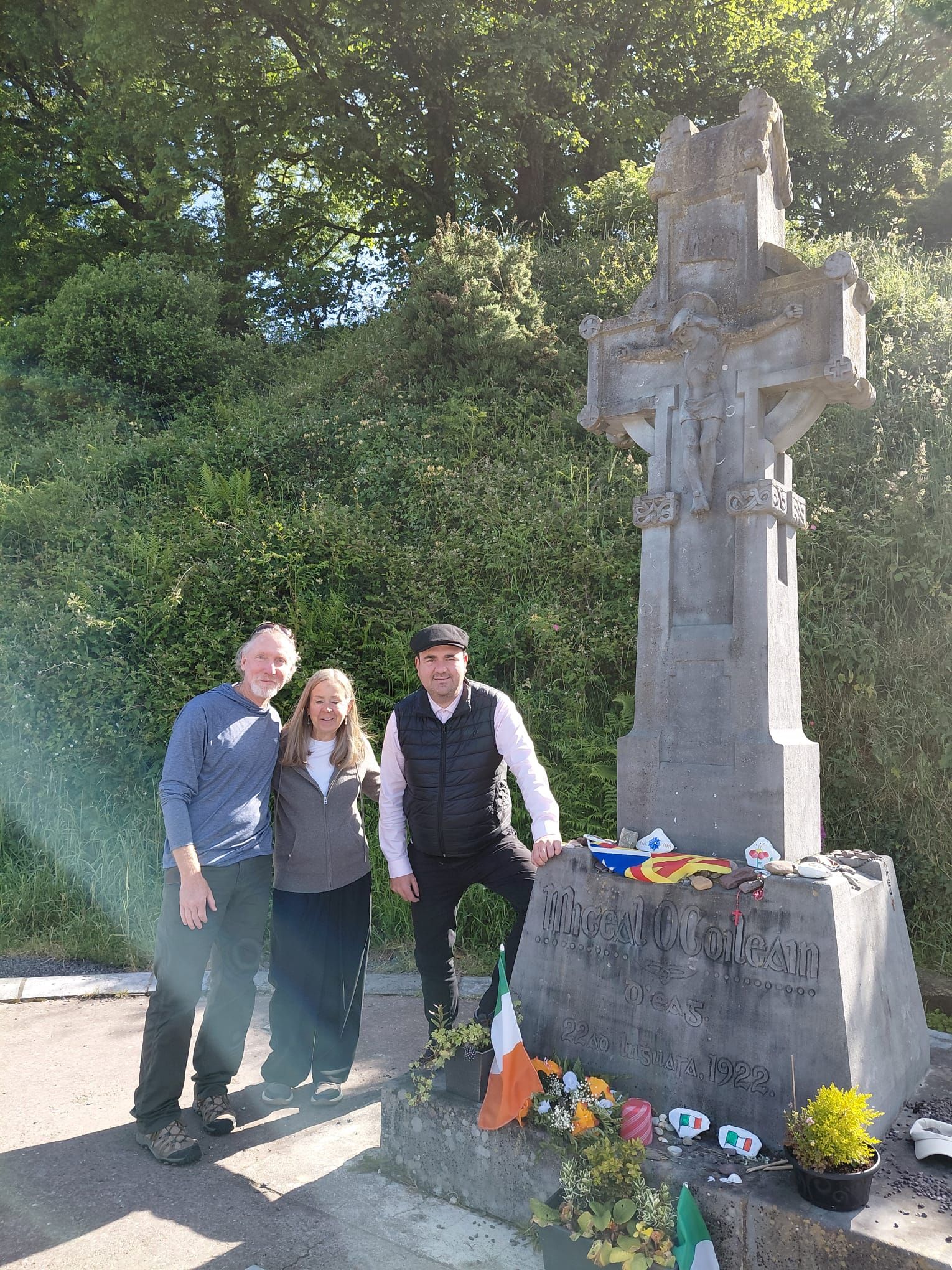 Two guests and their chauffeur stand beside a tall stone Celtic cross memorial on a sunny day.