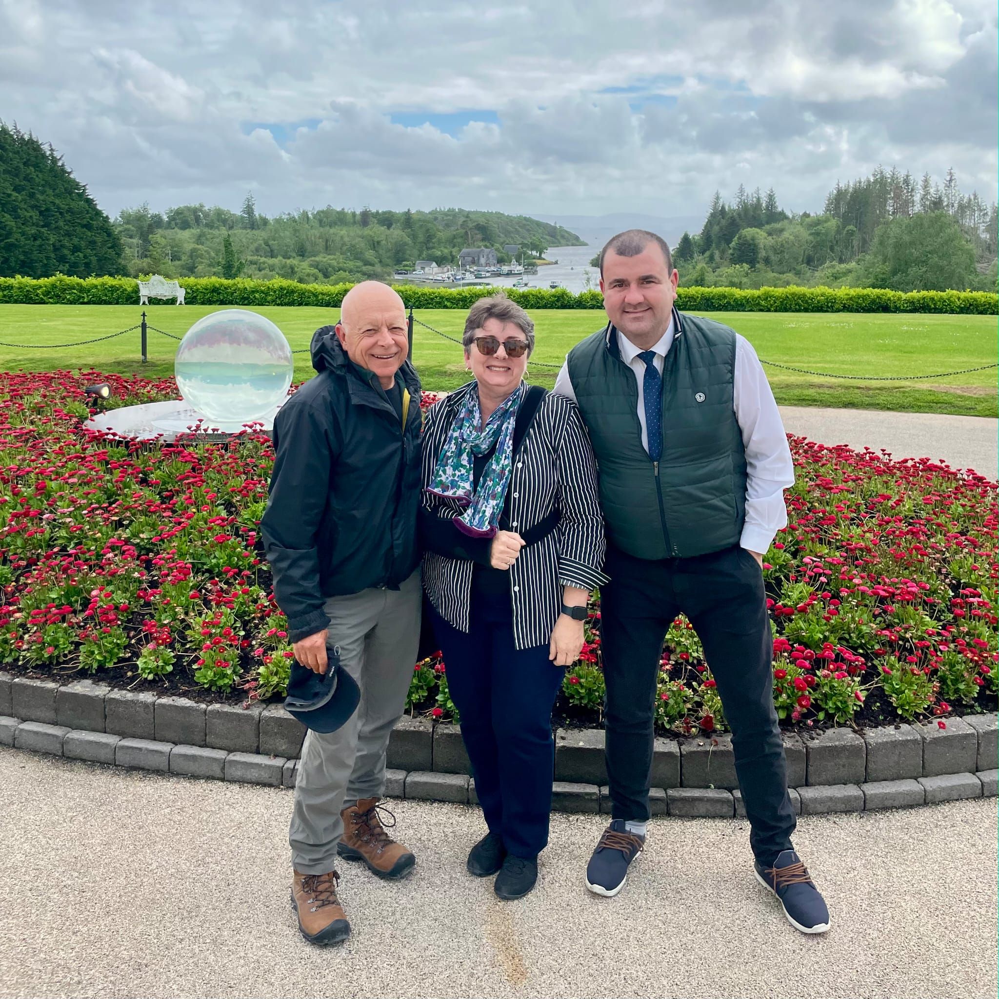 Two smiling guests stand with their chauffeur beside a red flower bed, with a lake and wooded hills behind them.