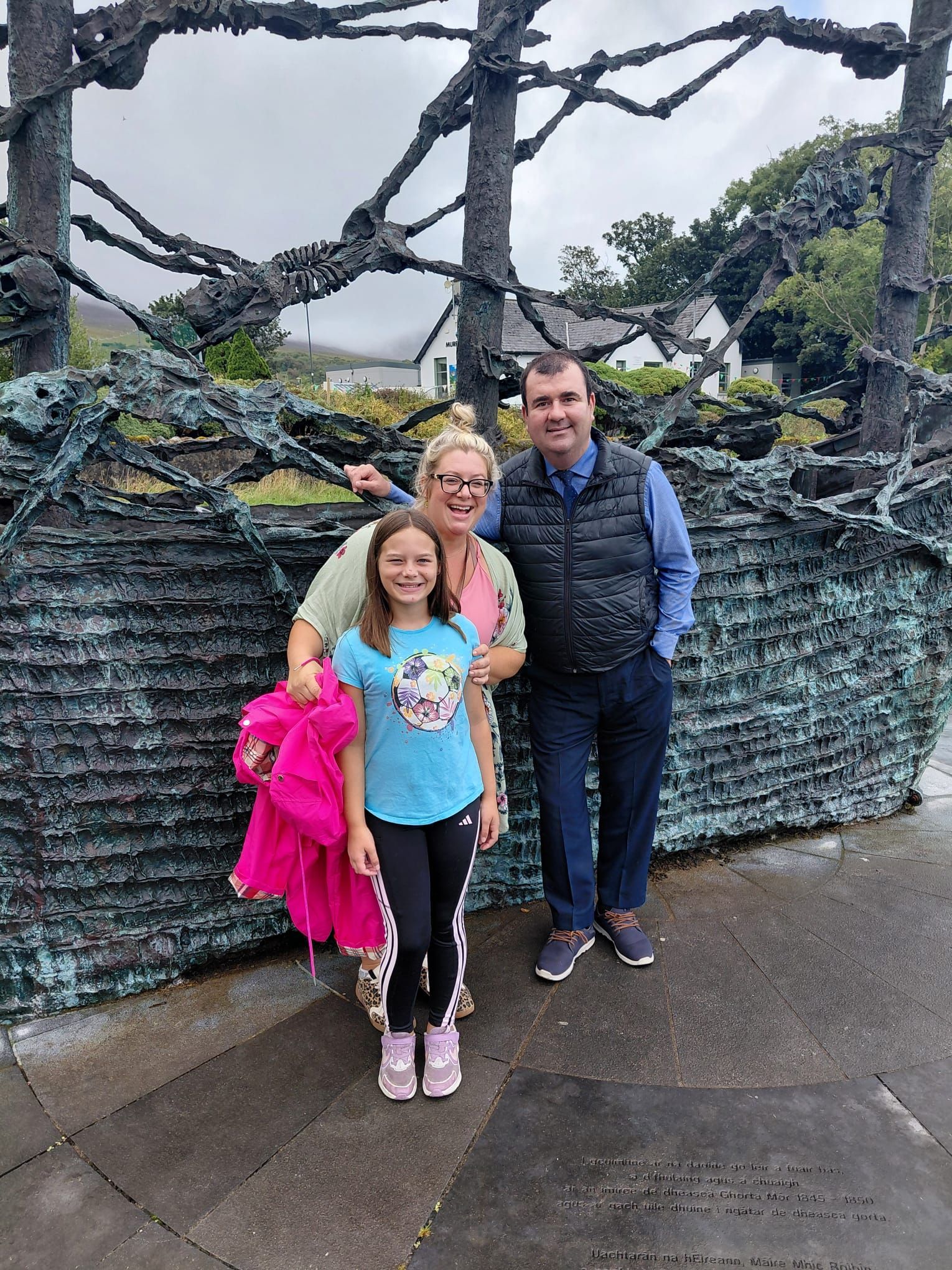 A mother, her daughter and their chauffeur smile in front of a large bronze sculpture of a ship.