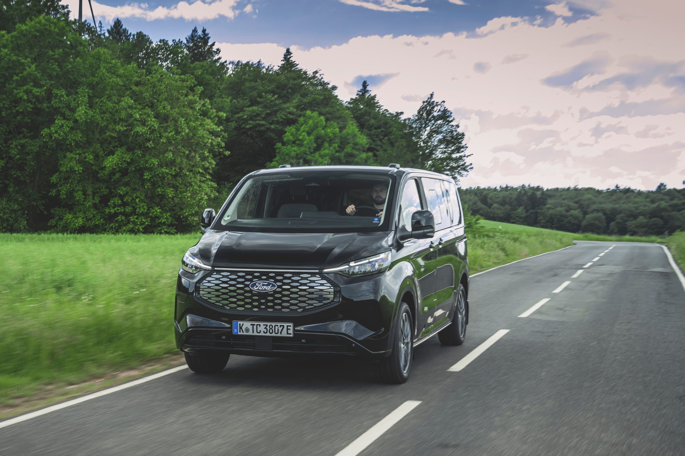 A black Ford Tourneo Custom drives along a country road between green fields and trees.