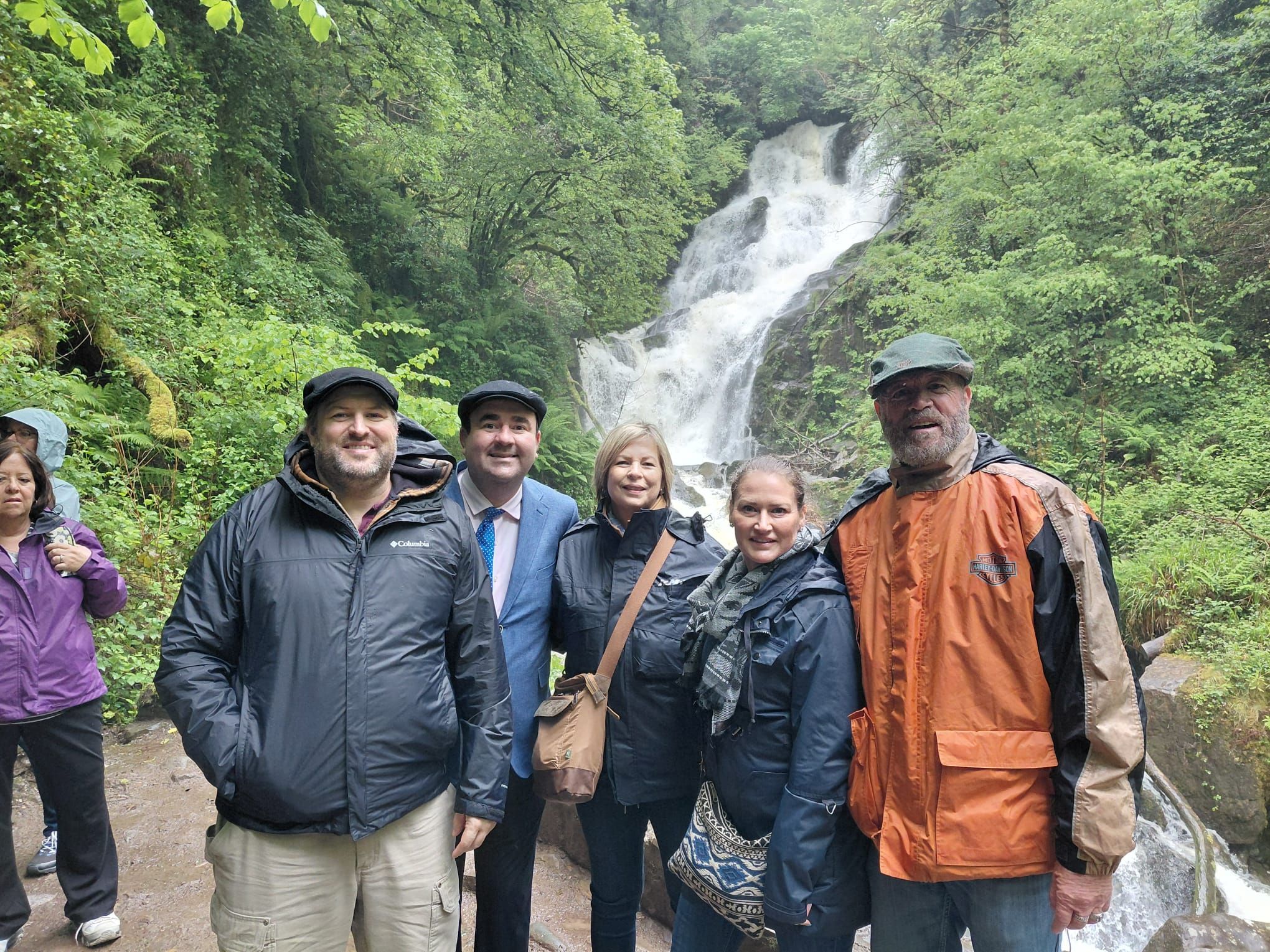 A group of guests in rain jackets smile in front of a waterfall in a green wooded glen.