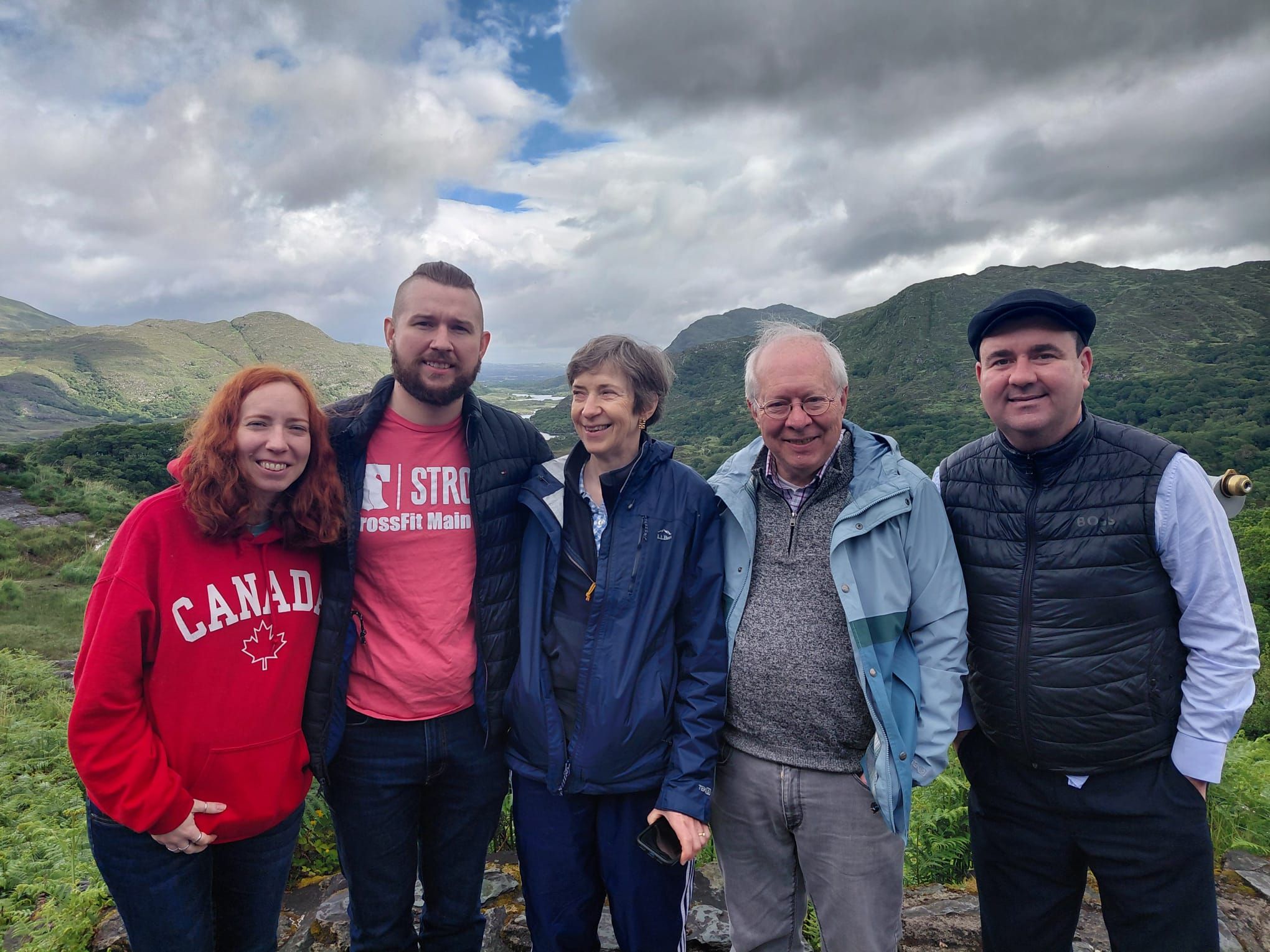 Paul stands with four guests at a viewpoint over a green valley with lakes and mountains under a cloudy sky.