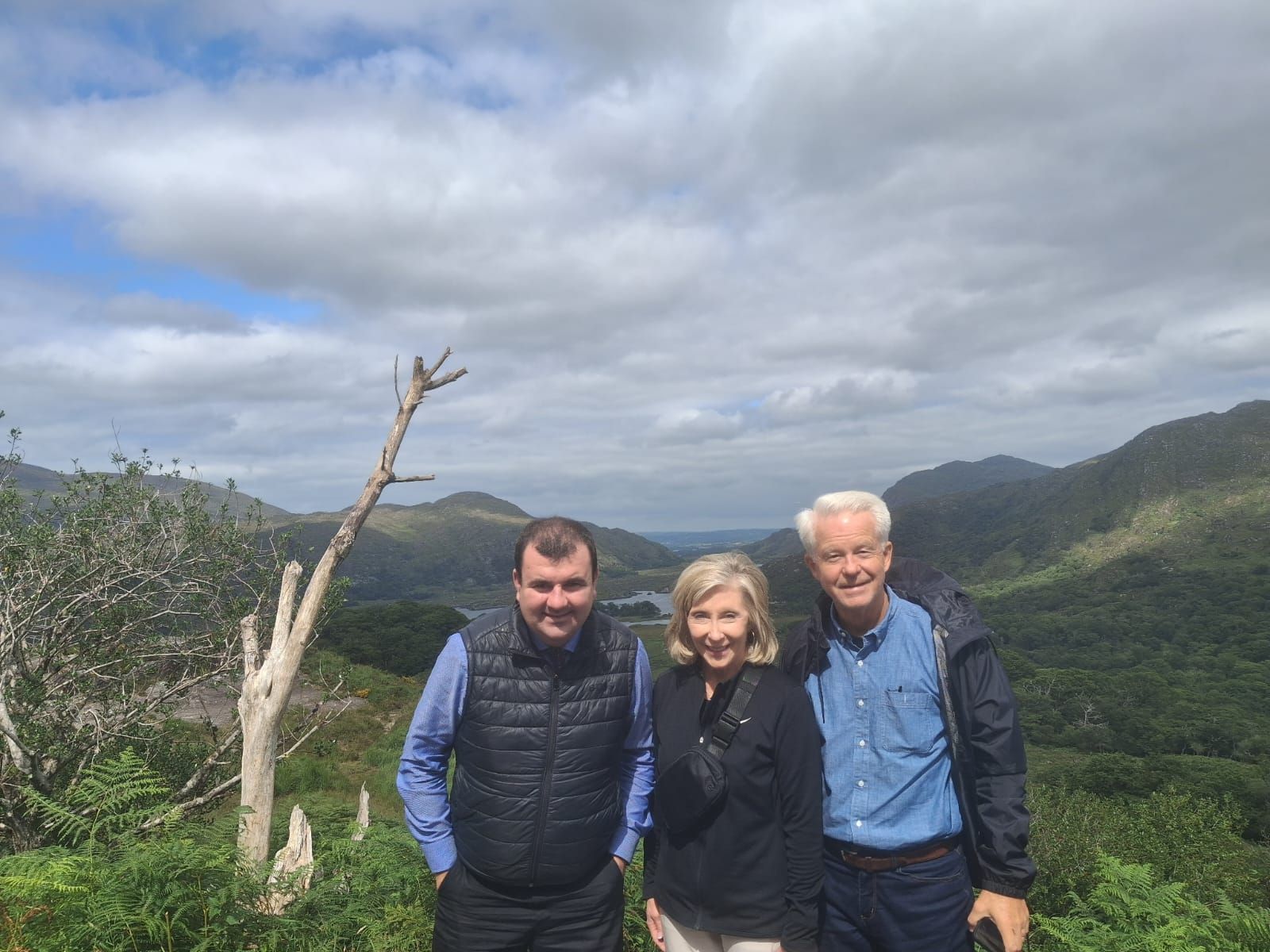 Paul and two guests stand at a viewpoint over wooded mountains and a lake, beside a bare dead tree.