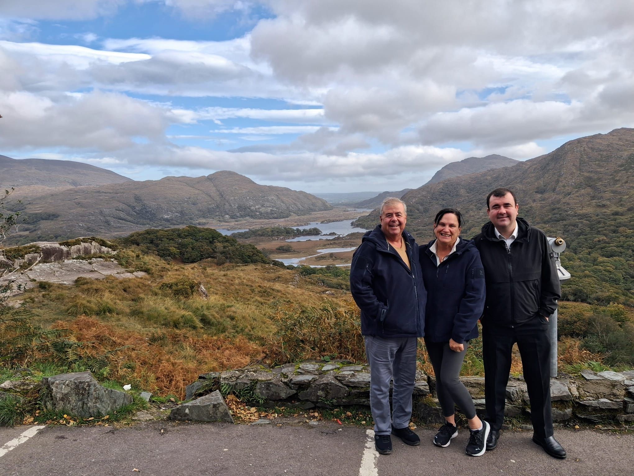 Paul and two guests stand at a stone wall above a valley of autumn bracken, lakes and mountains.
