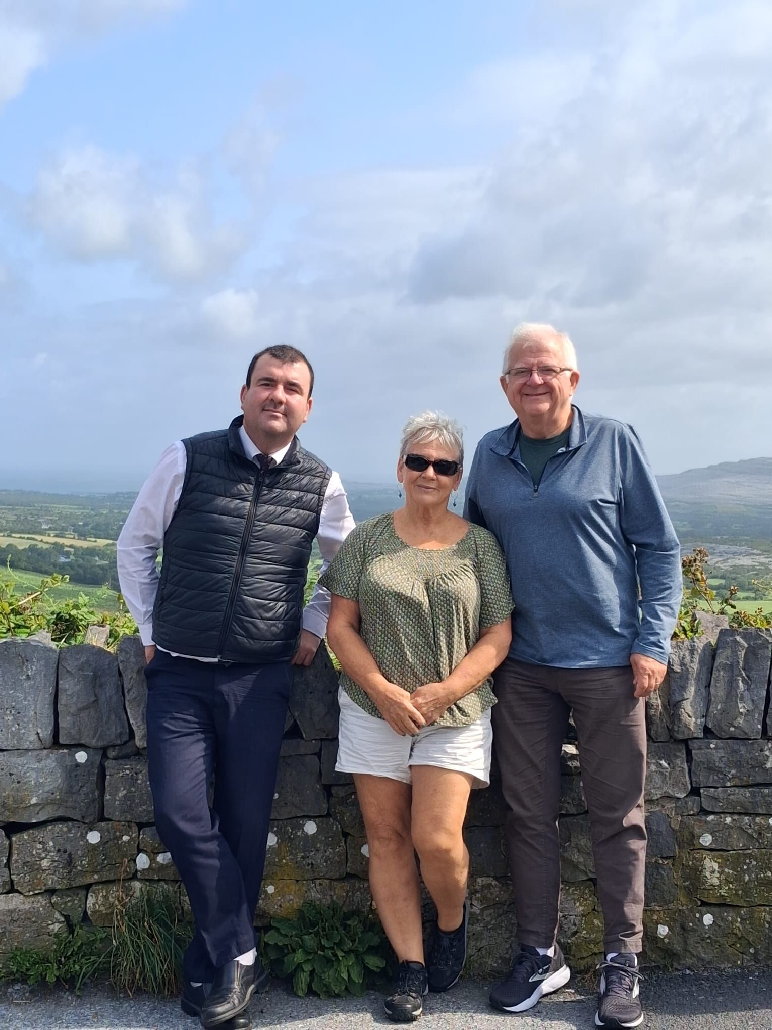 Paul and two guests lean on a dry-stone limestone wall, with green fields and hills behind.