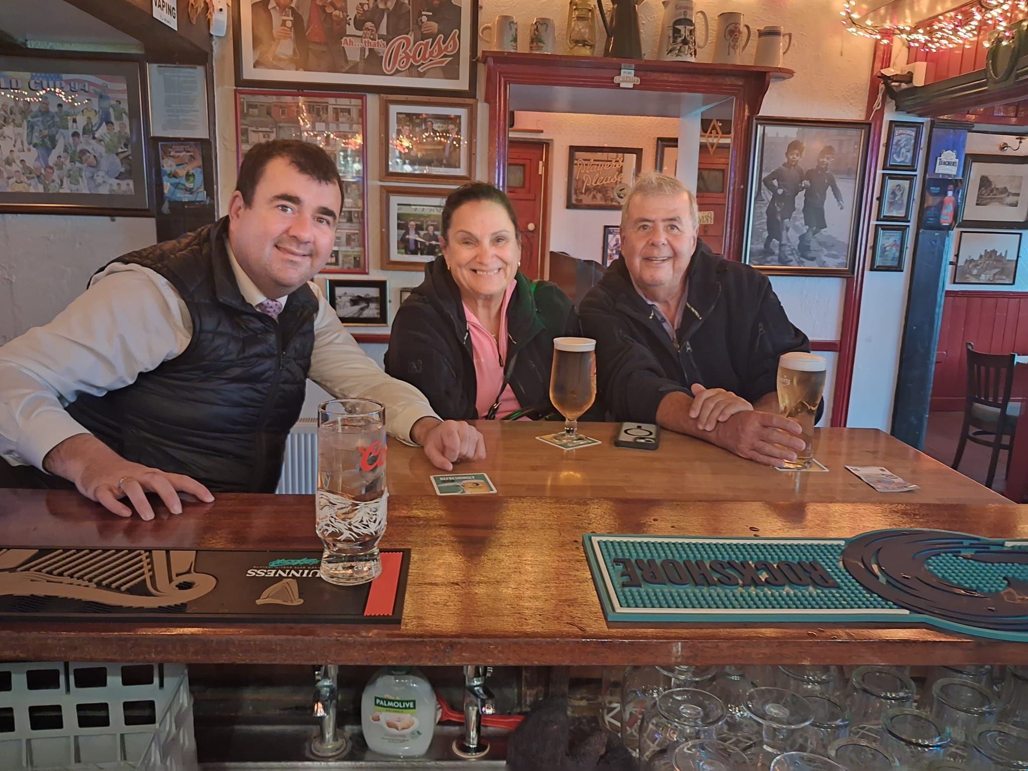 Paul and two guests smile at the bar of a traditional pub, with framed photos on the walls behind them.