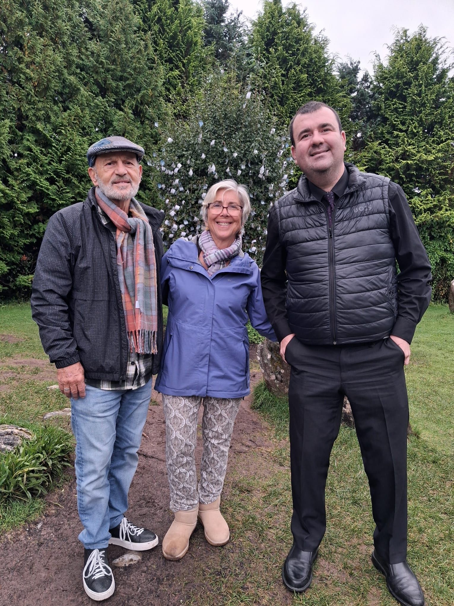 Paul stands with two guests on a grassy path in front of tall evergreen trees.
