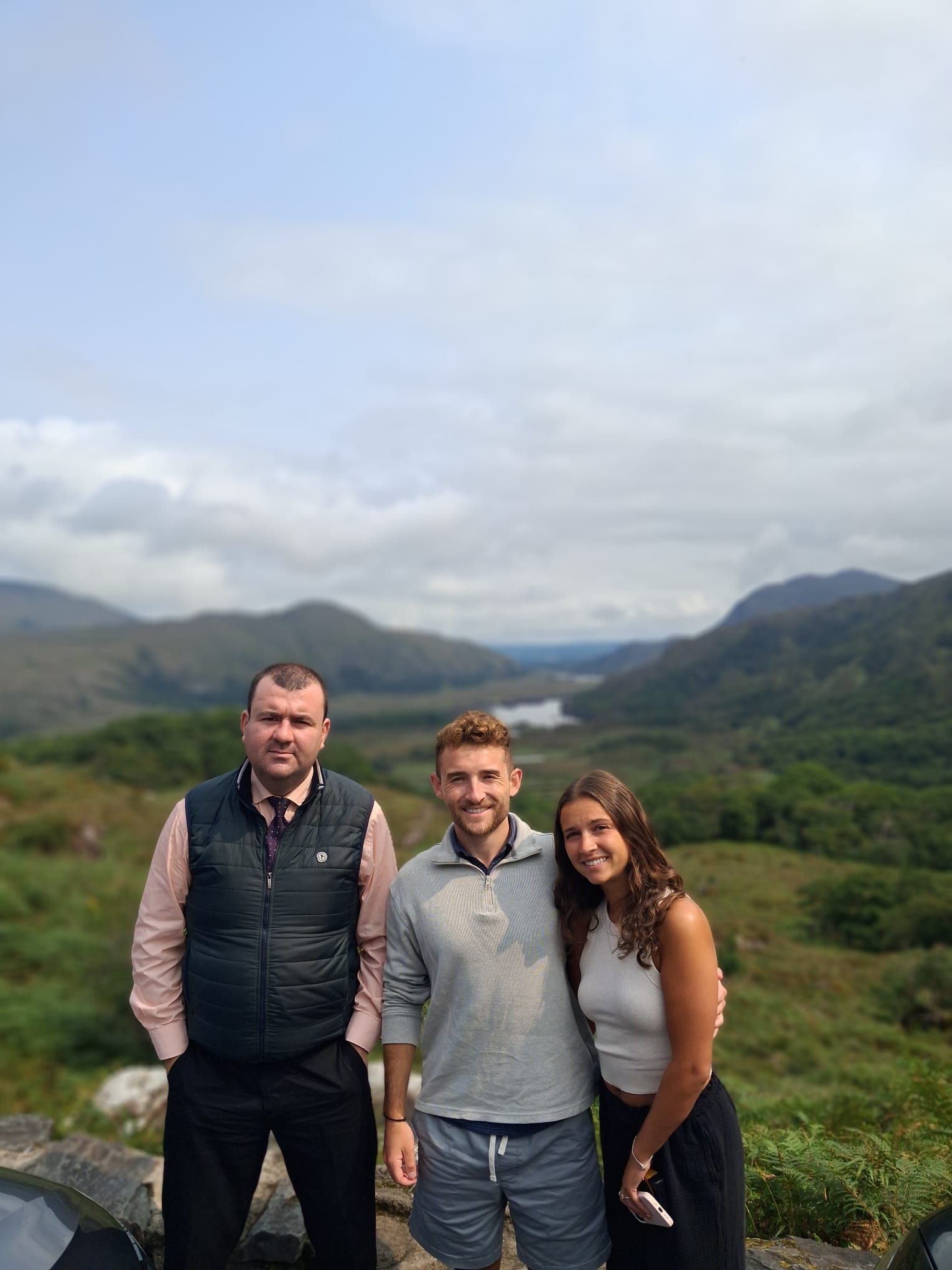 Paul stands beside a young couple, with a valley, a small lake and mountains far below.