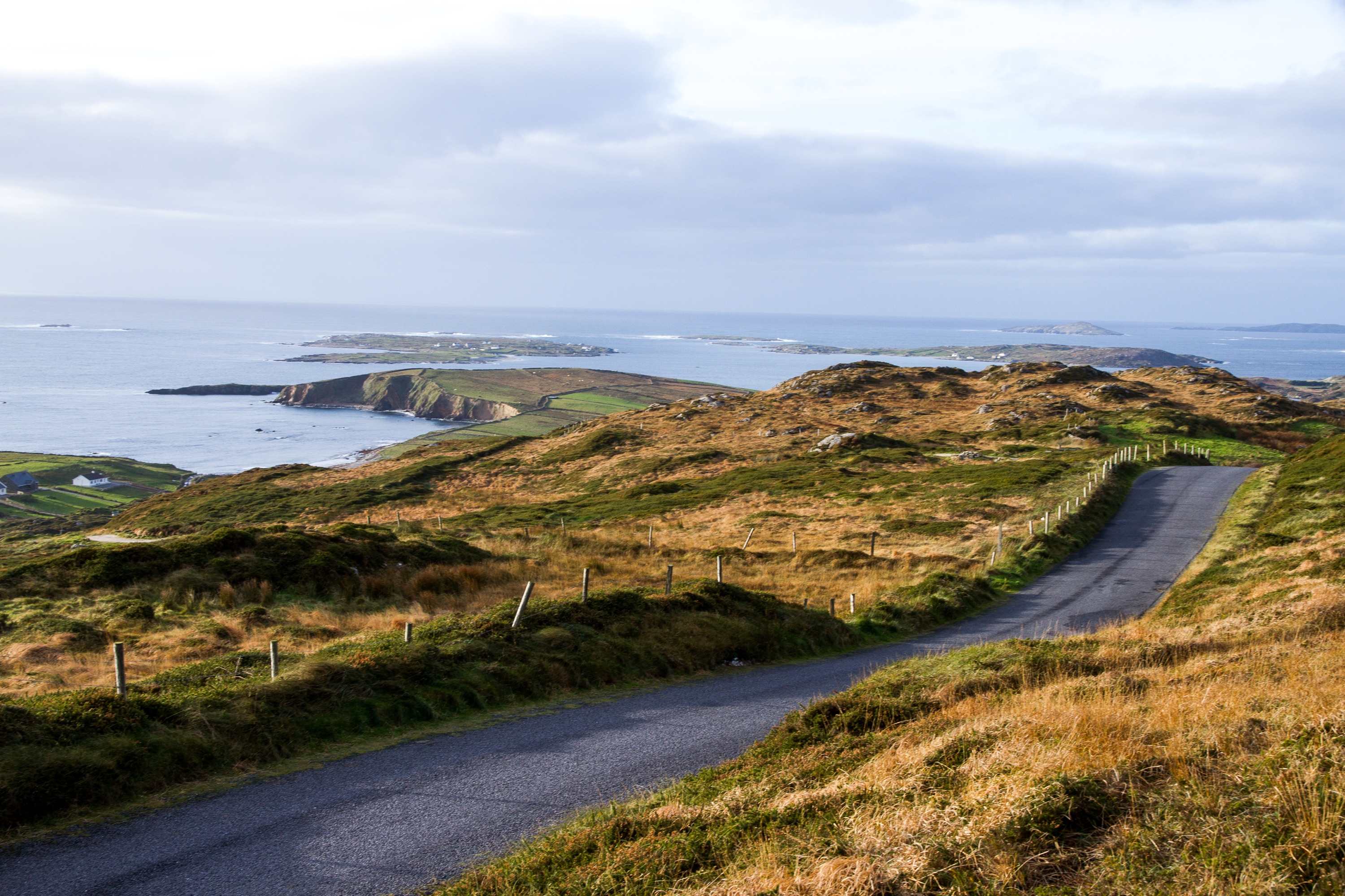 A narrow road winds over grassy hills, with the Atlantic, islands and headlands below under a pale sky.